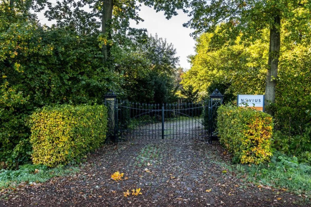 Zwarte toegangspoort tussen groene heggen bij een bosrijk pad aan de Langbroekerdijk, met een bord van Novius ernaast.